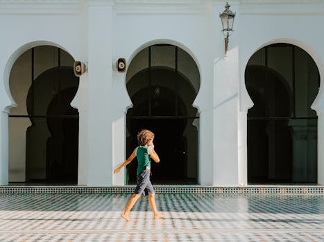 A child strolls in a sunlit Moroccan courtyard featuring traditional architecture with arches.