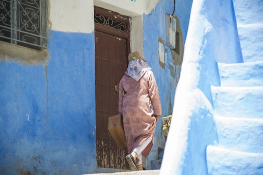 Full body back view of anonymous person wearing hat strolling with cardboard box along aged house with shabby door on street with staircase
