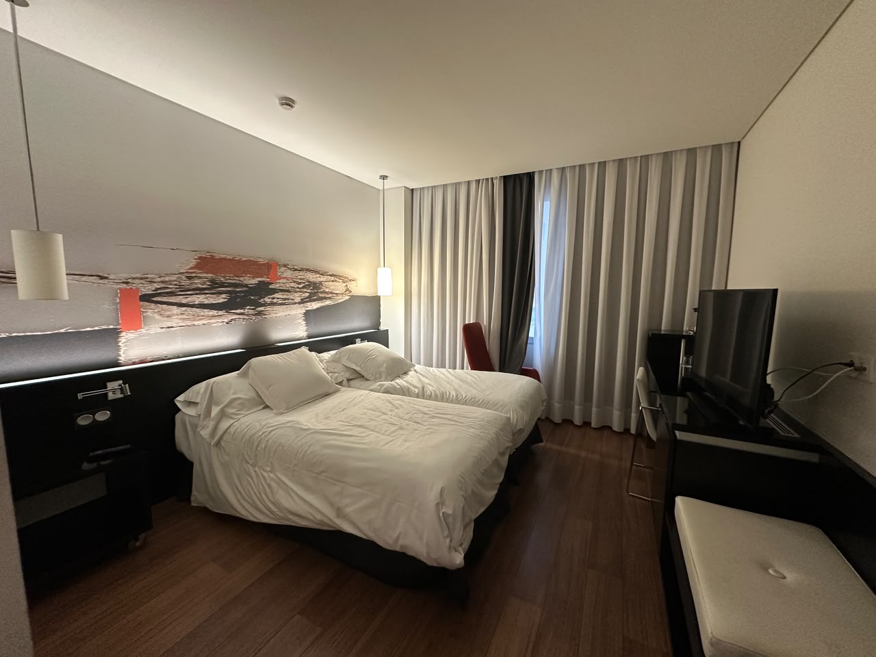Chambre meublée avec deux lits simples, un bureau, une télévision et de grands rideaux devant la fenêtre