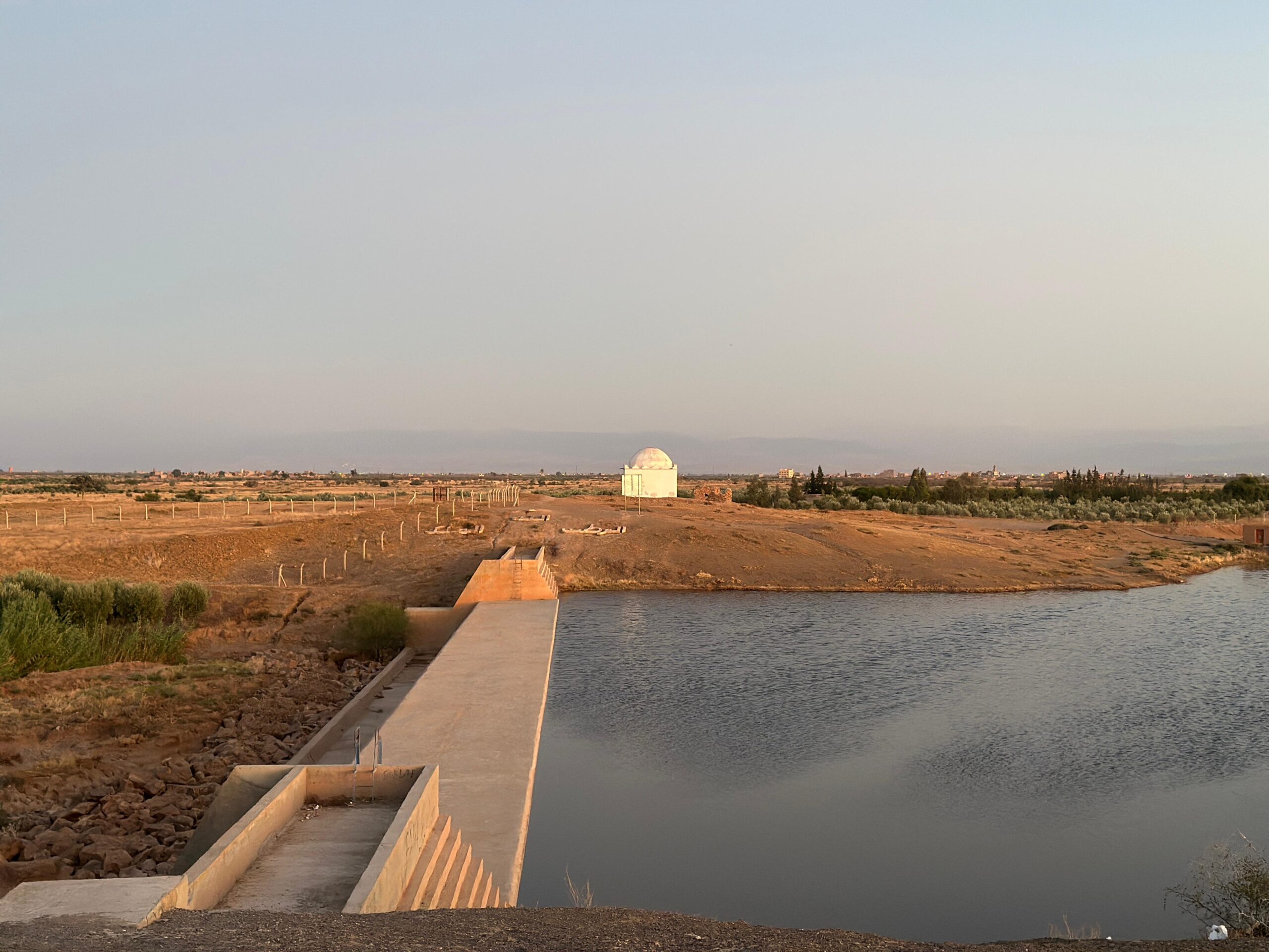 Small concrete reservoir in dry Moroccan countryside, with flat farmland and a white domed building on the horizon